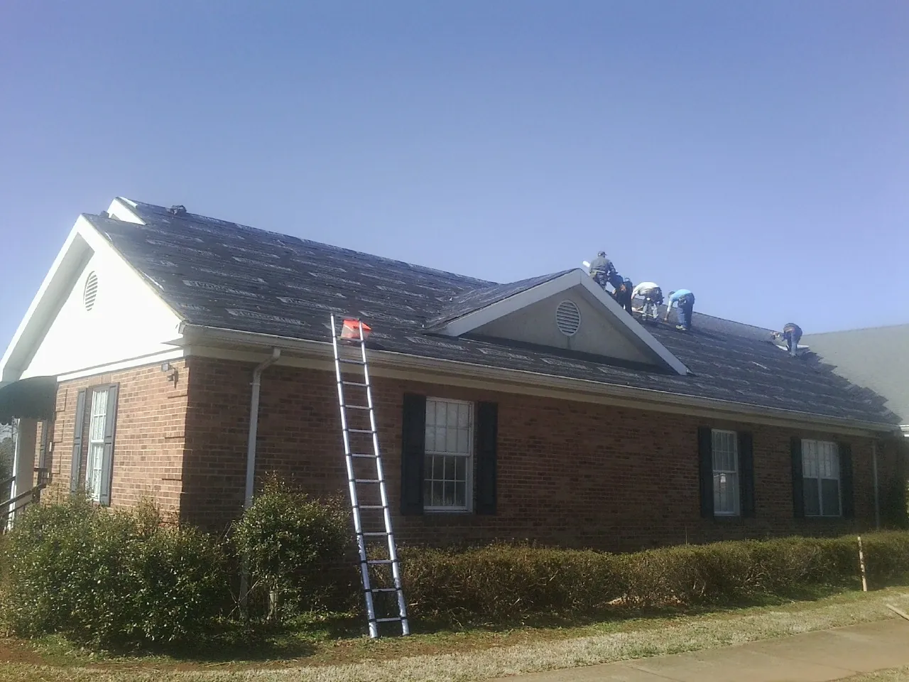 Crew tearing shingles off a brick ranch roof with a ladder set — Emergency Roof Repair work by our crew in Fort Mc Coy