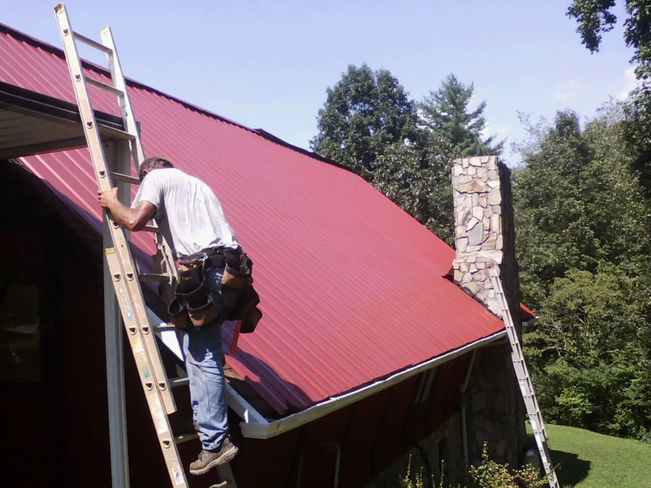 Roofer on a ladder beside a red metal roof and stone chimney — Soffit & Fascia Repair by Roofing Brothers in Fort Mc Coy